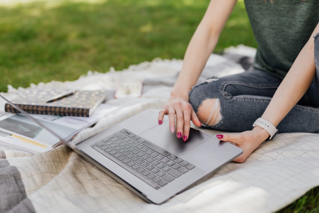 A woman working on a picnic blanket outside with laptop and notebook. Only the woman's lap and hands are in frame.